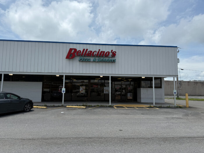 Exterior of Bellacino's Pizza & Grinders, a white metal-sided building with a red and green sign above the entrance. The front features a covered walkway and parking spaces directly in front.