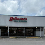Exterior of Bellacino's Pizza & Grinders, a white metal-sided building with a red and green sign above the entrance. The front features a covered walkway and parking spaces directly in front.