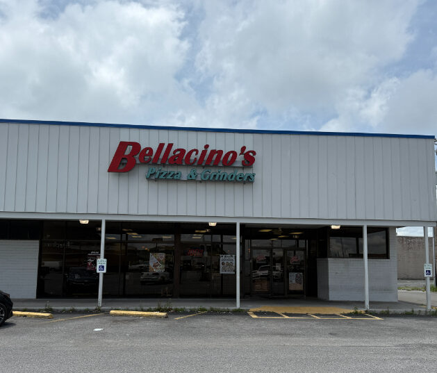 Exterior of Bellacino's Pizza & Grinders, a white metal-sided building with a red and green sign above the entrance. The front features a covered walkway and parking spaces directly in front.