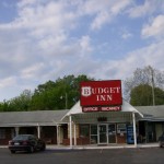 Exterior of the Budget Inn with a red sign above the entrance reading 'Budget Inn, Office, Vacancy,' and a single car parked in front.