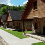 Row of rustic cabins at Buffalo Trail Cabins, featuring stone and wood exteriors, front porches with chairs, and a paved walkway, set against trees.