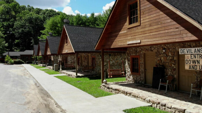 Row of rustic cabins at Buffalo Trail Cabins, featuring stone and wood exteriors, front porches with chairs, and a paved walkway, set against trees.