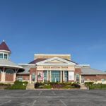 Front view of the Chuck Mathena Center, a large brick performing arts venue with large windows and columns, seen from the parking lot.
