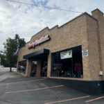 Exterior of Cole Harley-Davidson, a tan brick motorcycle dealership with large display windows, a black awning, and parking spaces in front along a roadside.