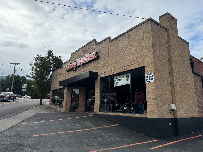 Exterior of Cole Harley-Davidson, a tan brick motorcycle dealership with large display windows, a black awning, and parking spaces in front along a roadside.