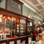 Interior of Bramwell Soda Fountain and Corner Shop, featuring antique wood cabinetry, chalkboard menus, rows of flavored syrups, and a staff member working behind the counter in a classic uniform. A vintage-style sign above reads "Welcome to the Corner Shop."