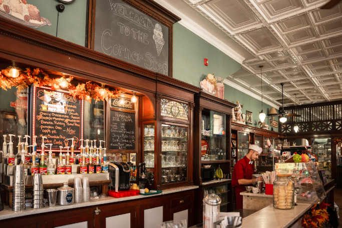 Interior of Bramwell Soda Fountain and Corner Shop, featuring antique wood cabinetry, chalkboard menus, rows of flavored syrups, and a staff member working behind the counter in a classic uniform. A vintage-style sign above reads "Welcome to the Corner Shop."