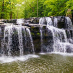 Wide, multi-tiered waterfall cascading over dark rock ledges into a calm pool below at Brush Creek Preserve and Falls, surrounded by lush green forest.