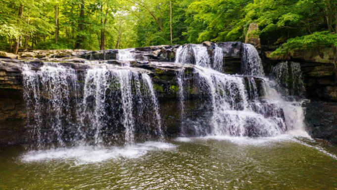 Wide, multi-tiered waterfall cascading over dark rock ledges into a calm pool below at Brush Creek Preserve and Falls, surrounded by lush green forest.