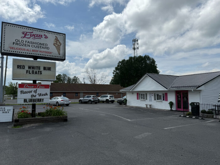 Exterior of Evan's Sweets, a white building with pink shutters and a large sign out front advertising frozen custard, hotdogs, and weekly flavors.