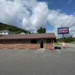 Front view of Farmer's Restaurant, a small brick building with a metal roof and a large roadside sign, set against a backdrop of hills and a partly cloudy sky.