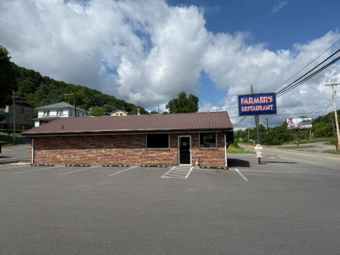 Front view of Farmer's Restaurant, a small brick building with a metal roof and a large roadside sign, set against a backdrop of hills and a partly cloudy sky.