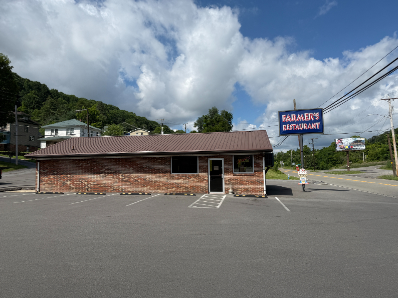 Front view of Farmer's Restaurant, a small brick building with a metal roof and a large roadside sign, set against a backdrop of hills and a partly cloudy sky.