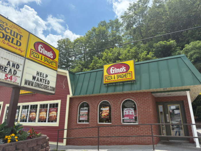 Exterior of Gino's Pizza & Spaghetti, a brick restaurant with a green roof and large yellow sign, next to a signboard advertising jobs.