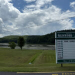 Glenwood Park entrance sign with a lake and tree-covered hills in the background under a partly cloudy sky.
