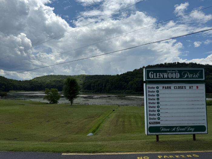 Glenwood Park entrance sign with a lake and tree-covered hills in the background under a partly cloudy sky.