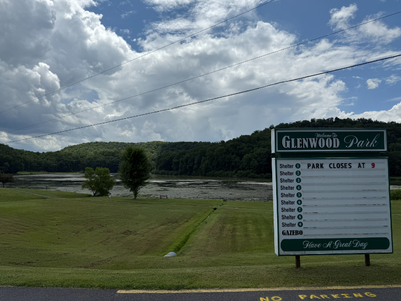 Glenwood Park entrance sign with a lake and tree-covered hills in the background under a partly cloudy sky.