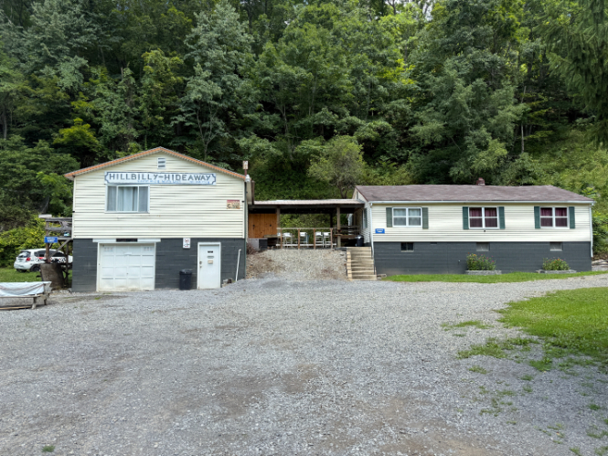 Hillbilly Hideaway lodging with two connected buildings, a gravel lot, and a forested hillside in the background.
