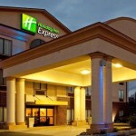 Exterior of the Holiday Inn Express in Princeton, illuminated at night with a large covered entrance supported by columns and the green and blue Holiday Inn Express logo lit on the building.