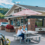 Lynn's, a classic drive-up restaurant with a brick exterior, striped umbrellas, and outdoor picnic tables. Two people are seated at a table enjoying their food, with cars parked nearby and flower planters out front.