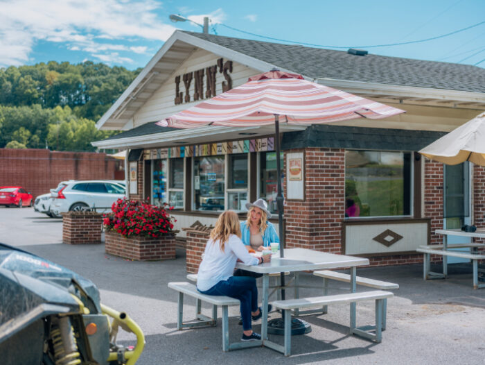 Lynn's, a classic drive-up restaurant with a brick exterior, striped umbrellas, and outdoor picnic tables. Two people are seated at a table enjoying their food, with cars parked nearby and flower planters out front.
