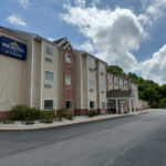Exterior of the Microtel Inn by Wyndham in Princeton, a three-story beige and brick building with a peaked entrance and a large roadside sign. The driveway wraps around the front, with parked cars and landscaped shrubs lining the edge.