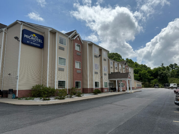 Exterior of the Microtel Inn by Wyndham in Princeton, a three-story beige and brick building with a peaked entrance and a large roadside sign. The driveway wraps around the front, with parked cars and landscaped shrubs lining the edge.