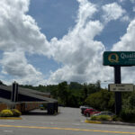 Exterior of the Quality Inn in Princeton, with a tall roadside sign and a single-story brick building set back from the road. A driveway leads to a parking lot lined with trees and cars.