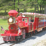 The Ridge Runner, a bright red miniature train ride with passengers, traveling on a gravel track through a wooded park area.