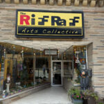 Front of The RiffRaff Arts Collective, a storefront with large display windows, a colorful sign, and potted plants by the entrance. A carved bear statue stands to the right of the door.