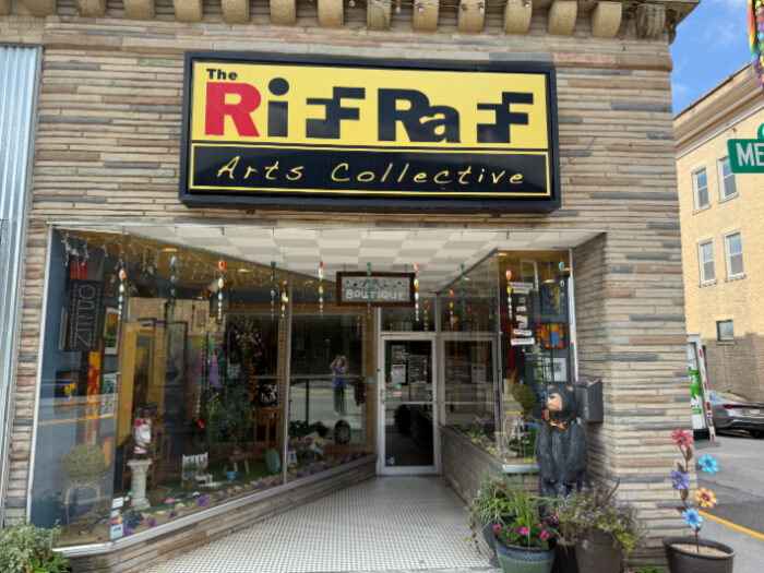 Front of The RiffRaff Arts Collective, a storefront with large display windows, a colorful sign, and potted plants by the entrance. A carved bear statue stands to the right of the door.