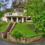 Two-story cream-colored house with a large front porch at River View Inn LLC, situated on a raised lawn with stone steps and a retaining wall, surrounded by trees.