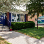 Exterior of Sisters Coffee House, a brick building with dark blue colors, potted plants on the steps, and a sign out front featuring a blue teacup and mountain design.