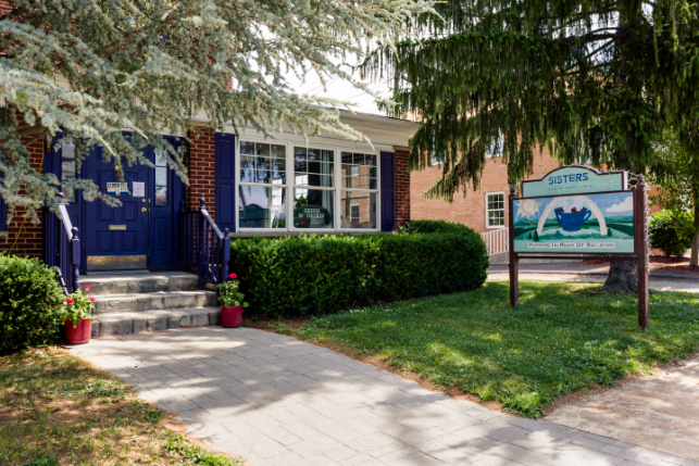 Exterior of Sisters Coffee House, a brick building with dark blue colors, potted plants on the steps, and a sign out front featuring a blue teacup and mountain design.