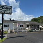 Exterior of Valley Country Restaurant, a white building with a covered front porch and patio seating, featuring a large roadside sign with the restaurant's name and phone number.