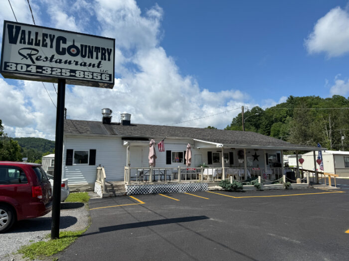 Exterior of Valley Country Restaurant, a white building with a covered front porch and patio seating, featuring a large roadside sign with the restaurant's name and phone number.
