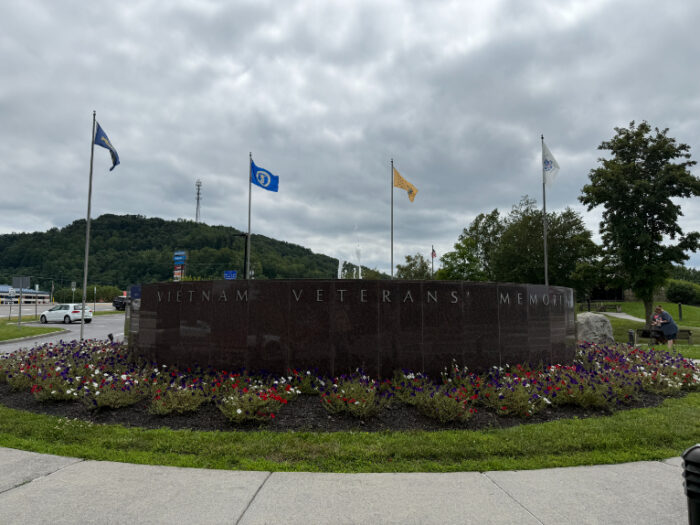 Outdoor Vietnam Veterans Memorial made of stone with engraved text, surrounded by colorful flower beds and flagpoles flying various flags, set against a cloudy sky.