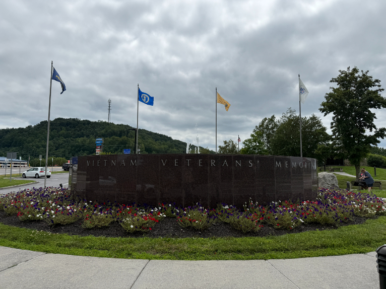Outdoor Vietnam Veterans Memorial made of stone with engraved text, surrounded by colorful flower beds and flagpoles flying various flags, set against a cloudy sky.