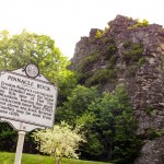 Historic marker sign in front of a tall rocky cliff surrounded by greenery at Pinnacle Rock State Park, with the stone formation rising prominently in the background.