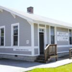 Front view of the Mercer County Agricultural Museum, a small gray building with white trim and a sign on the window displaying the museum's name. A simple wooden ramp and stairs lead to the entrance.