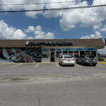 Front of Cheap Thrills Records, a music store with a dragon and lightning mural on the exterior wall, a row of display windows, and cars parked in the lot under a partly cloudy sky.