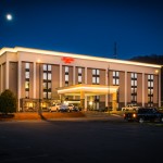 Exterior of the Hampton Inn in Princeton, shown at night with the building lit up and a well-lit entrance canopy. The multi-story hotel features tall windows, a red Hampton Inn logo, and cars parked out front.