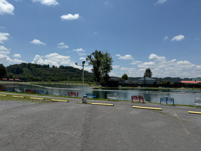A peaceful fishing pond with colorful benches along the edge, surrounded by wooded hills and a blue sky.