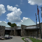 Stone building with triangular glass roof sections, three flagpoles out front, and a sign near the entrance, home to WV Crafts and Gifts.