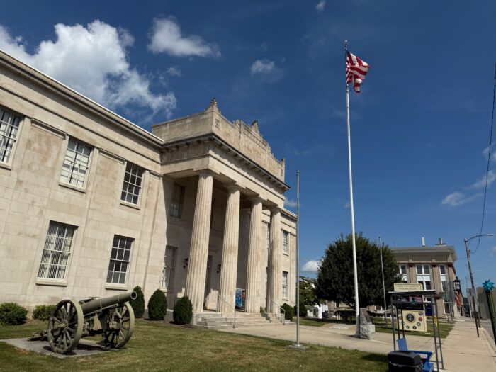 Stone government building with tall columns, an American flag on a pole, and a historic cannon on the lawn in front of the Mercer County War Museum in Princeton, West Virginia