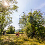 Overgrown amusement park ride surrounded by trees and tall grass at Lake Shawnee Abandoned Amusement Park, with a few visitors walking nearby under a sunny sky.