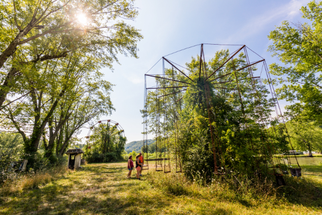 Overgrown amusement park ride surrounded by trees and tall grass at Lake Shawnee Abandoned Amusement Park, with a few visitors walking nearby under a sunny sky.