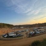 Dirt racetrack at sunset with cars, trucks, and trailers in the infield.