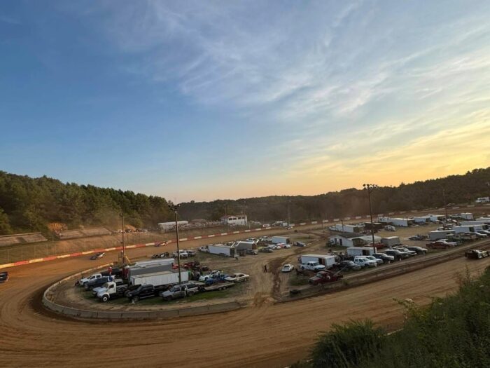 Dirt racetrack at sunset with cars, trucks, and trailers in the infield.