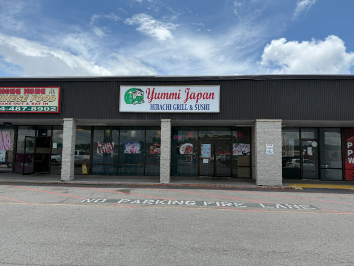 Front of Yummi Japan Hibachi Grill & Sushi, located in a shopping plaza with a white sign and red lettering above the entrance. Large glass windows display photos of menu items.